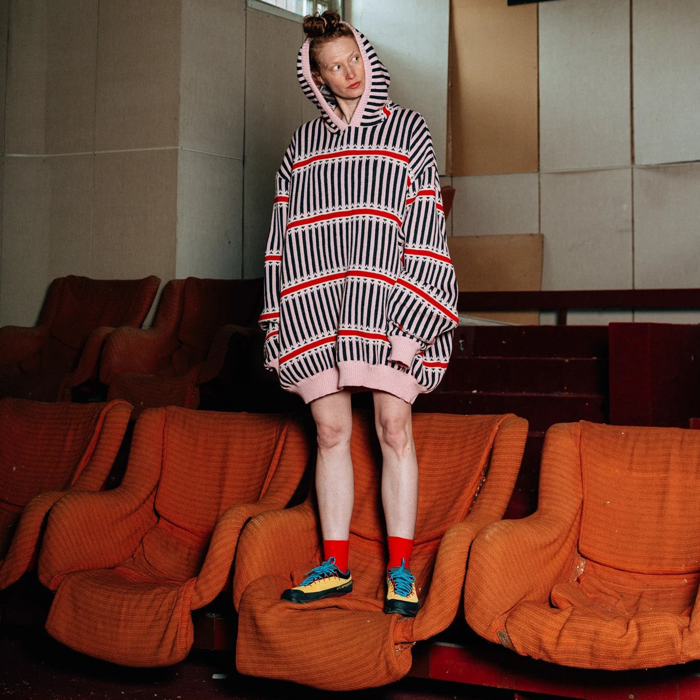 Person wearing a striped hoodie in an abandoned theater with orange seats and a tiled wall.
