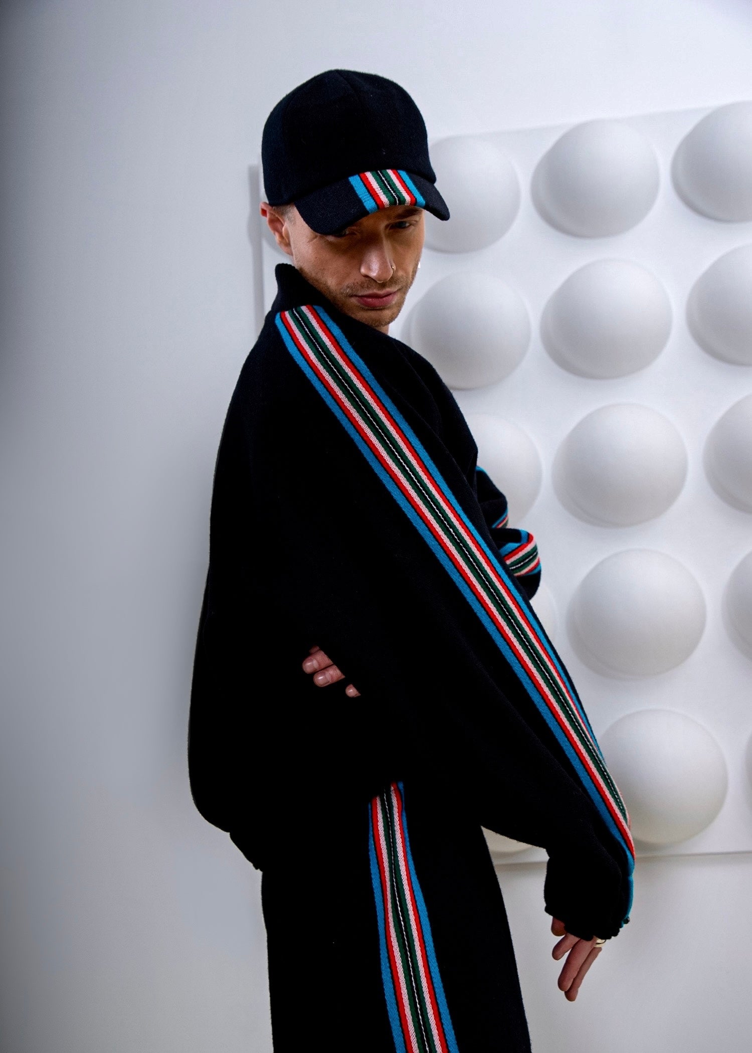 Person wearing a black jacket with colorful stripes and a maching cap, standing against a white textured wall.