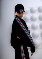 Person wearing a black jacket with colorful stripes and a maching cap, standing against a white textured wall.