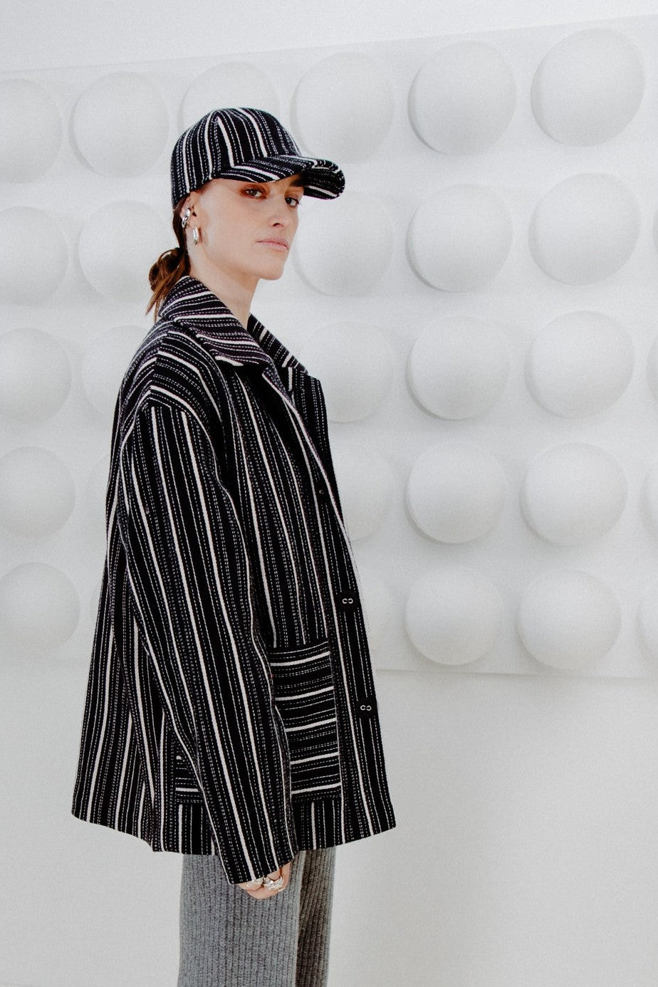 Person wearing a black wool jacket with white stripes and a maching cap, standing against a white textured wall.