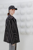 Person wearing a black wool jacket with white stripes and a maching cap, standing against a white textured wall.